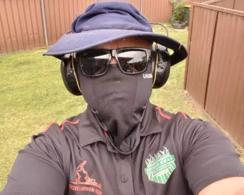 A man in a hat and sunglasses taking a selfie while mowing the lawn.