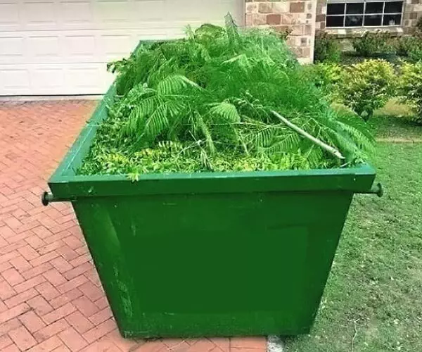 A green trash can filled with plants in front of a house offering Garden Services.